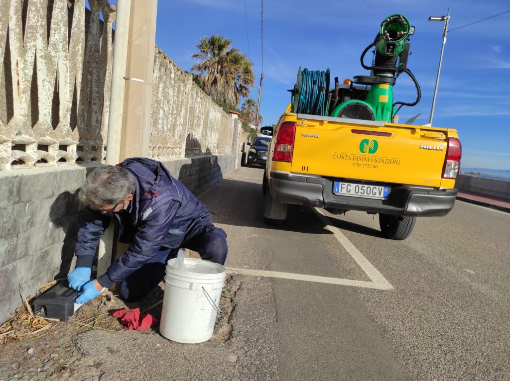 Operatore con guanti blu posiziona un erogatore per esche accanto a un muro, dietro un pick-up giallo Costa Disinfestazioni